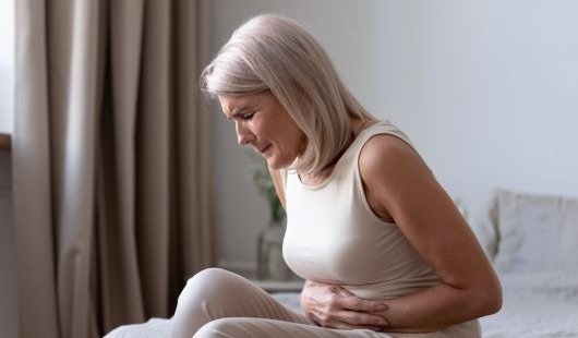 A woman in pain sitting on her bed clutching her stomach. 